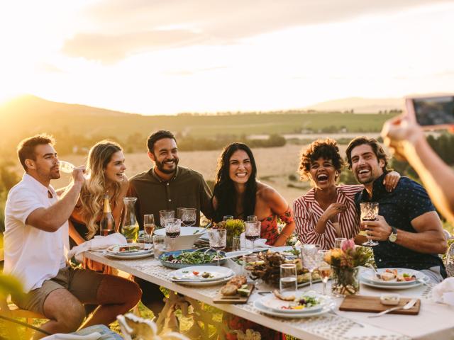 Group of men and women posing for a photograph during an outdoor dinner party. Woman hands taking picture of her friends at dinner party.