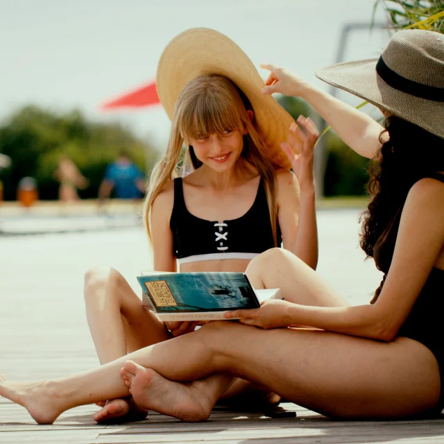 une fille et une femme assises au bord de la piscine en maillot de bain, chapeau et livre à la main.
